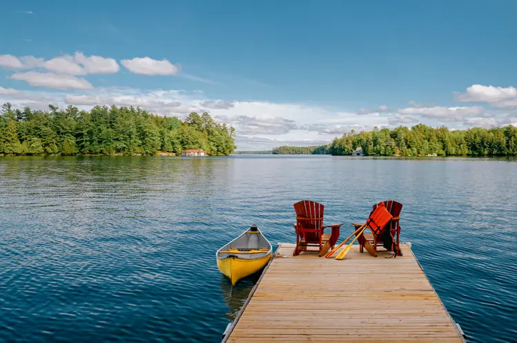 Un muelle de madera con sillas Adirondack y una canoa amarilla, con vista a un lago tranquilo y una costa boscosa bajo un cielo azul claro.
