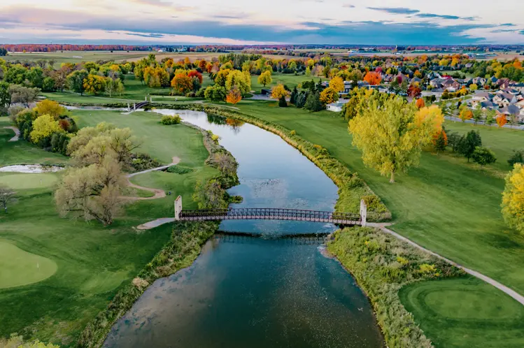 골프장을 지나 흐르는 강, 보행자 다리, 그리고 지평선 너머로 보이는 멀리 보이는 마을의 생동감 넘치는 가을 풍경입니다.
