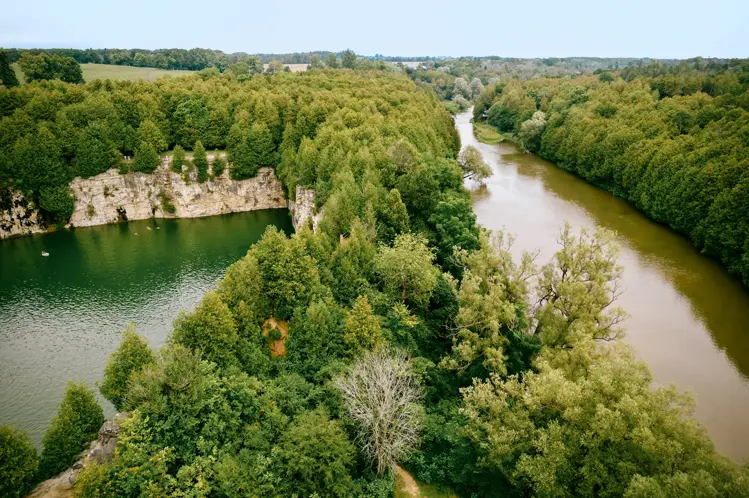 Draufsicht auf eine bewaldete Landschaft mit einem smaragdgrünen Steinbruchsee und einem gewundenen Fluss.