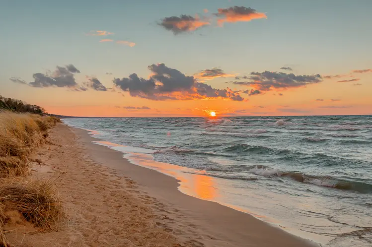 A sandy beach along Lake Huron at sunset, with the sun reflecting on the water and waves gently rolling ashore under a colourful sky.