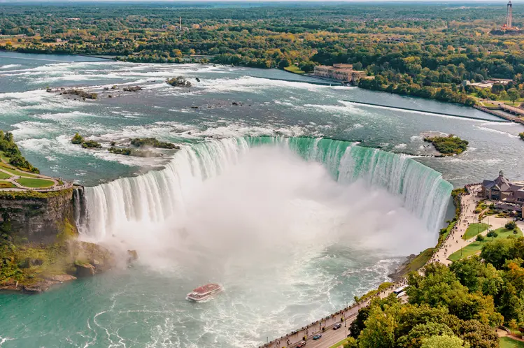 Vue aérienne des chutes Horseshoe du côté canadien avec un bateau touristique en contrebas et de la brume s'élevant sous un ciel bleu.