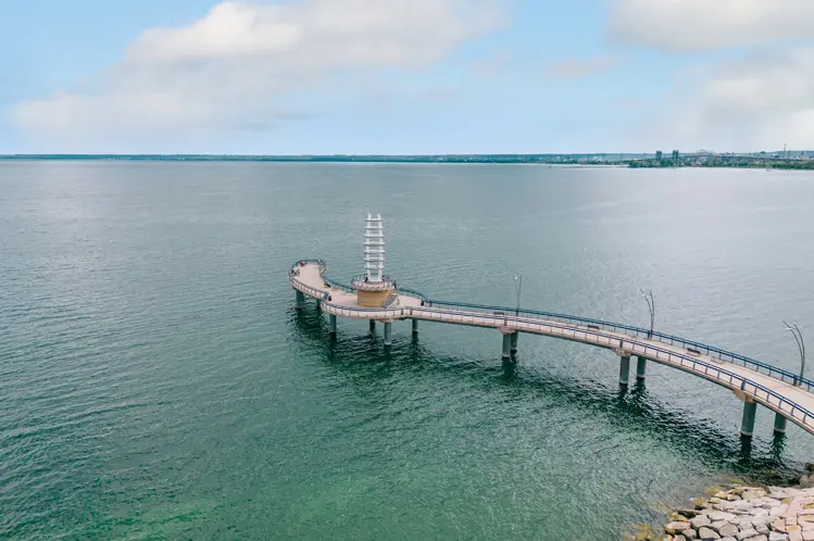  흐린 하늘 아래 온타리오 호수의 청록색 바닷물에 브랜트 스트리트 부두가 굽이쳐 흐르고, 방문객들이 부두를 따라 걷고 있습니다.