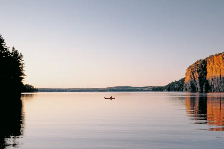 A person canoeing across a calm lake at sunset in Bon Echo Provincial Park, with forest on one side and a rocky cliff reflecting in the water.