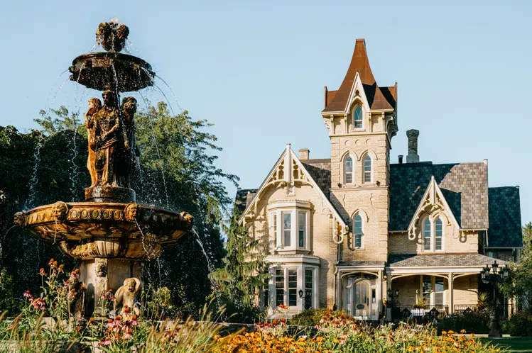 Victorian Gothic-style Elm Hurst Inn & Spa, with a sculpted tiered fountain and vibrant garden blooms under a clear blue sky.