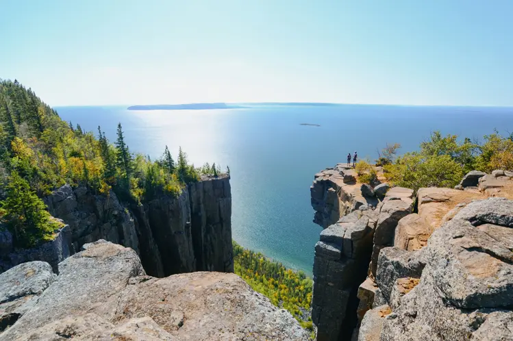 Two hikers standing on a rocky cliff overlooking Lake Superior at Sleeping Giant Provincial Park on the Sibley Peninsula, under a bright blue sky