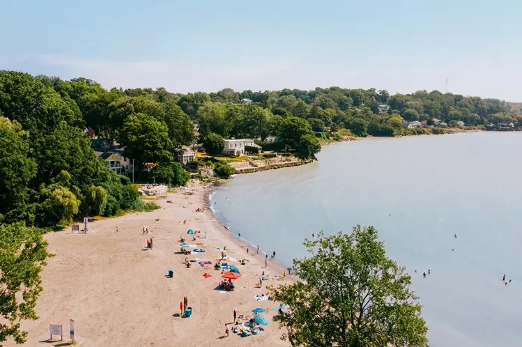 Beachgoers relaxing along the sandy shoreline of Lake Erie in Port Stanley, with calm waves and cottages nestled among lush trees.