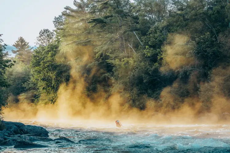  Remador navegando por rápidos brumosos en el río Madawaska en las tierras altas de Haliburton, rodeado de un denso bosque y luz de la mañana temprano.