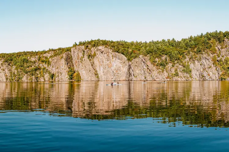 Towering Mazinaw Rock reflecting in Upper Mazinaw Lake, framed by forest and sandy shoreline at Bon Echo Provincial Park.