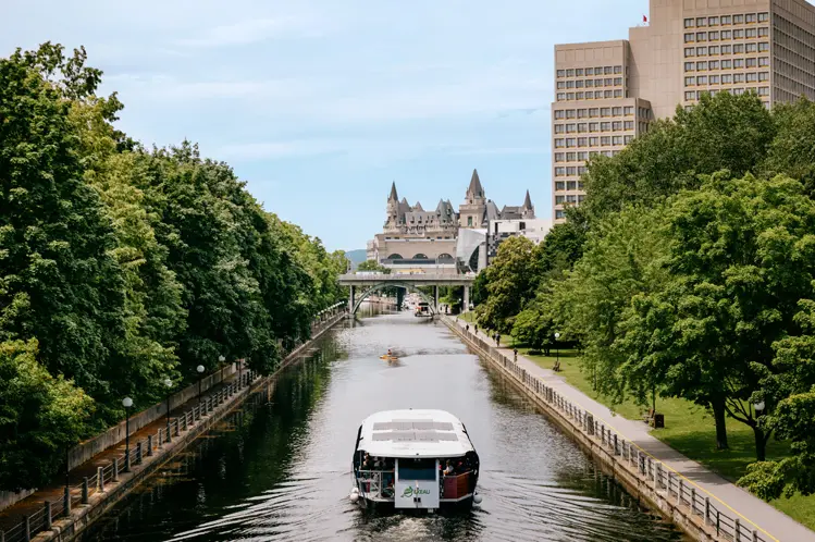 A boat cruising down the tree-lined Rideau Canal in Ottawa, with Parliament and a high-rise visible in the distance.