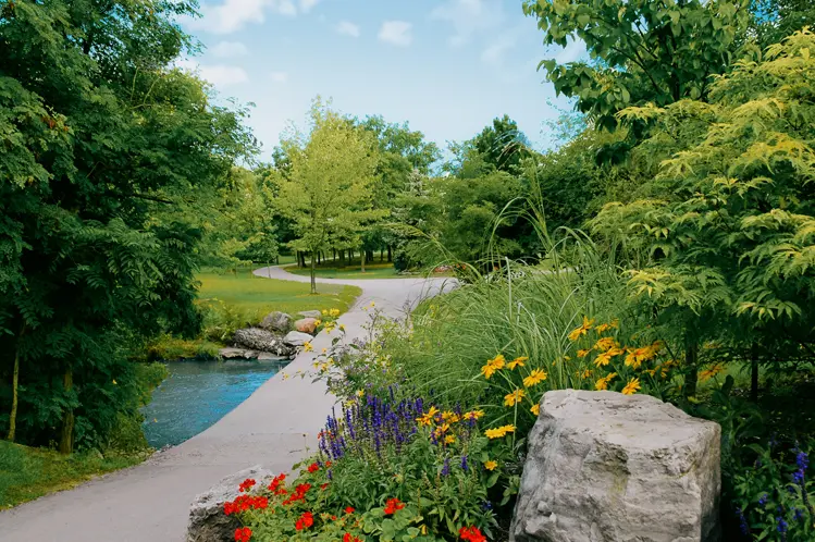 Scenic walking path in Brampton lined with summer flowers, trees, and a creek under a bright blue sky.