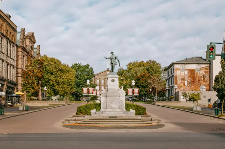 Historic downtown Brockville with a central war memorial, heritage buildings, and tree-lined streets under a cloudy sky.