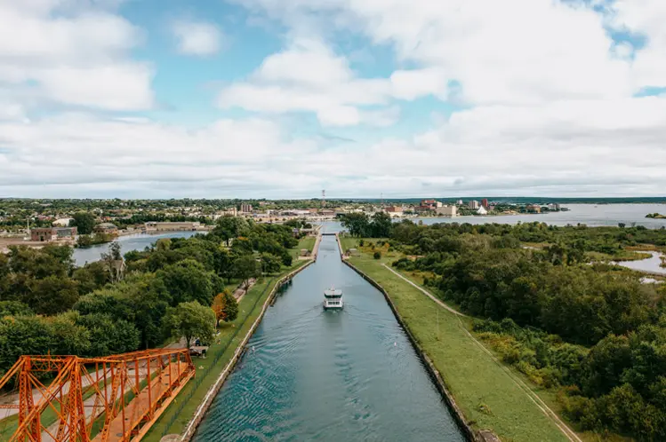 A boat cruising through the Sault Ste. Marie Canal on a cloudy summer day, passing beneath a red swing bridge with greenery along the shoreline.