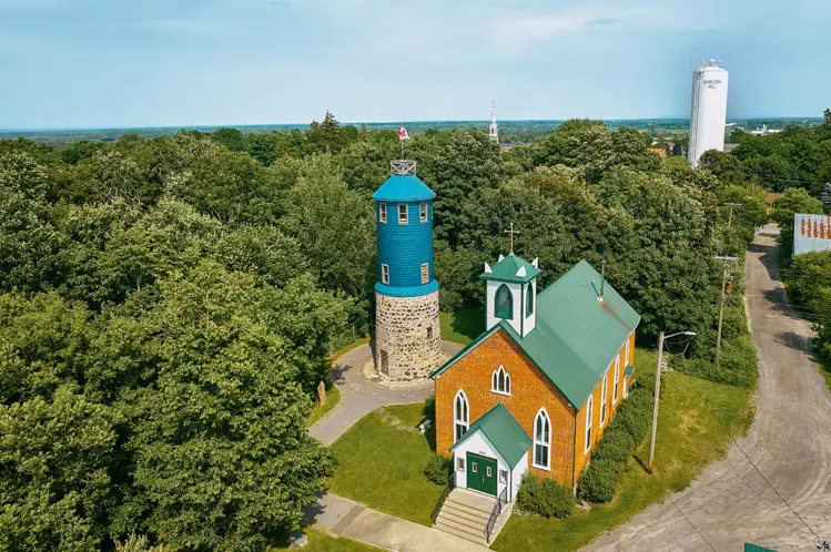 Vista aérea de la Torre Higginson y la Iglesia Anglicana de San Juan el Apóstol en Vankleek Hill, rodeada de árboles verdes en un día de verano.