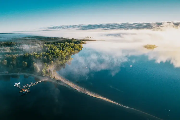 Vue aérienne du parc provincial Wabakimi, avec un hydravion amarré, une forêt dense et un lac brumeux, immobile et vitreux au lever du soleil.