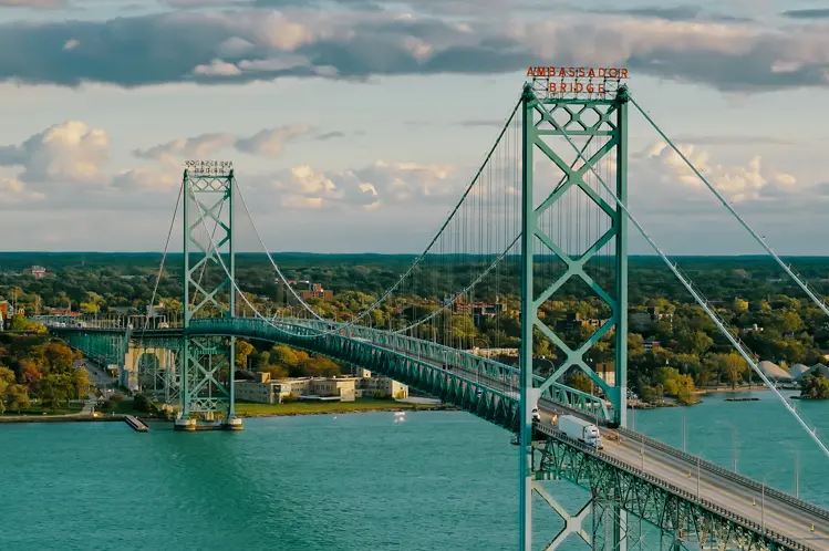 Aerial view of the Ambassador Bridge stretching across turquoise waters, connecting Windsor, Ontario, with the U.S., under a sunset sky.