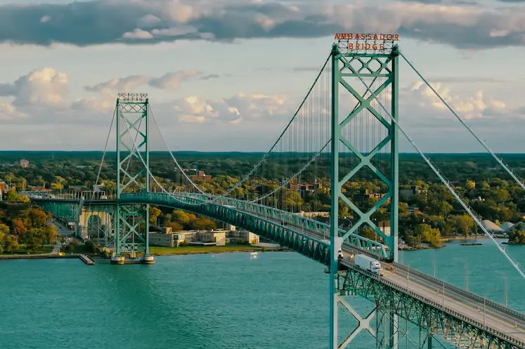 Aerial view of the Ambassador Bridge stretching across turquoise waters, connecting Windsor, Ontario, with the U.S., under a sunset sky.