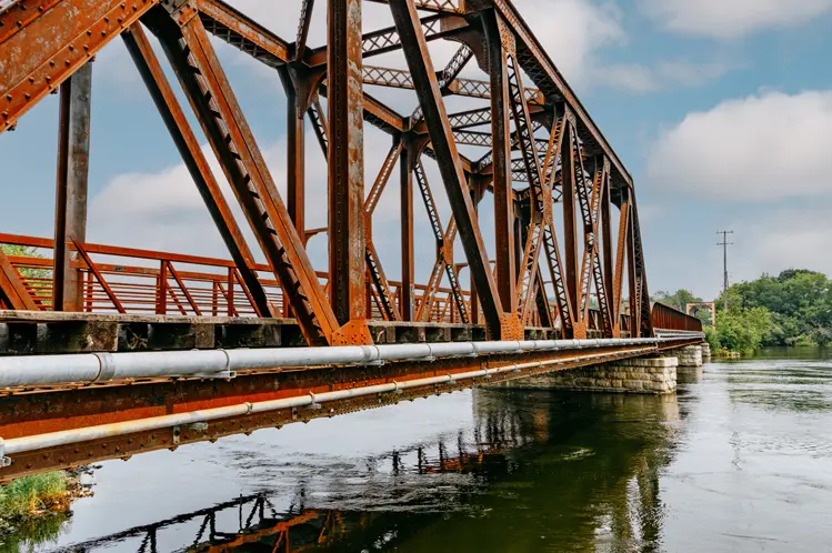 Puente ferroviario de color óxido sobre el río Otonabee a lo largo del Sendero Transcanadiense en Peterborough, reflejado en aguas tranquilas.