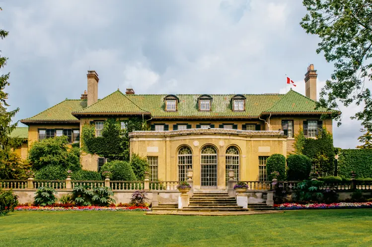 Rear view of Parkwood Estate mansion, featuring a grand stone facade, arched windows, green roof and manicured gardens under a cloudy summer sky.