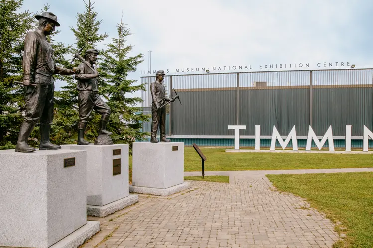 Mining statues outside the Timmins Museum National Exhibition Centre, with large TIMMINS letters in the background on a cloudy day.