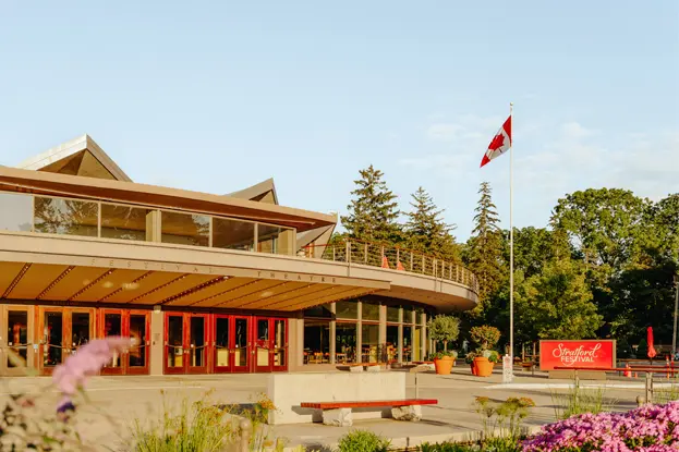  여름의 스트랫퍼드 페스티벌 극장. 붉은 문, 캐나다 국기가 펄럭이고, 맑은 하늘 아래 정원이 꽃으로 뒤덮여 있습니다.