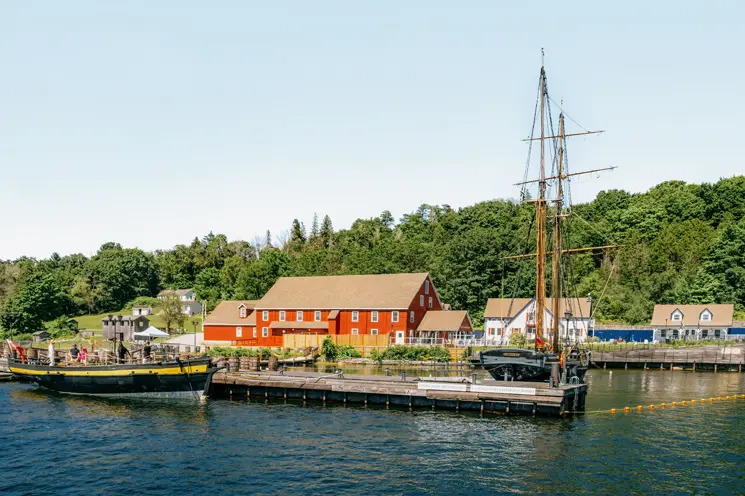 Un gran velero atracado en Discovery Harbour en Penetanguishene, Ontario, con edificios patrimoniales de color rojo y una exuberante vegetación bajo un cielo claro de verano.