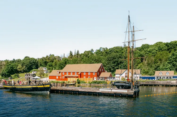 Grand voilier amarré au port Discovery à Penetanguishene, en Ontario, avec des bâtiments patrimoniaux et une végétation luxuriante sous un ciel bleu.