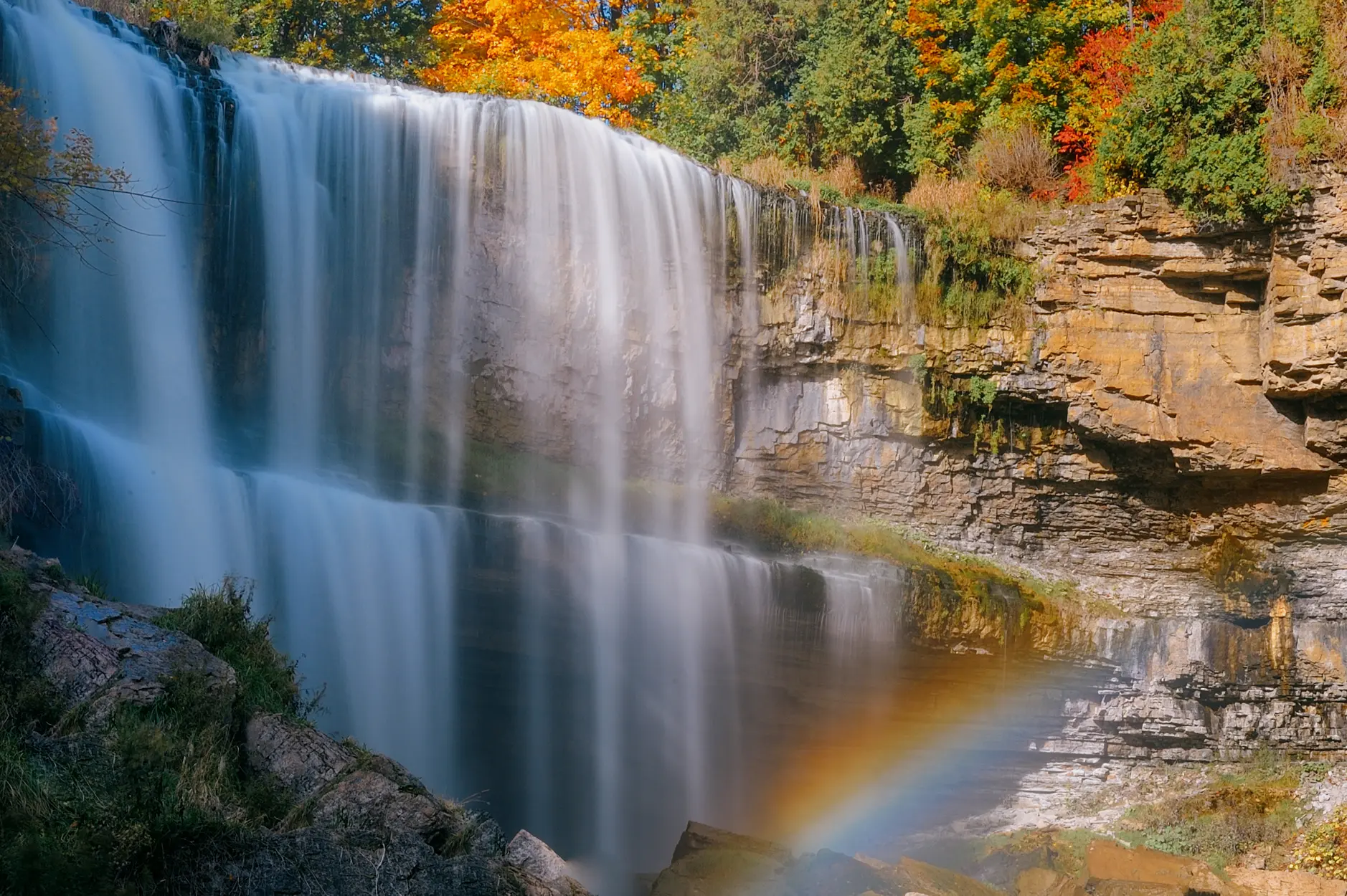 A cascading waterfall over the rocky escarpment surrounded by fall foliage and a rainbow in the foreground.