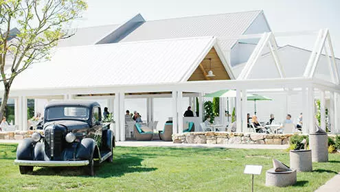 A black vintage car sits on the grass in front of a large white open air gazebo filled with people sitting on chairs and drinking wine.