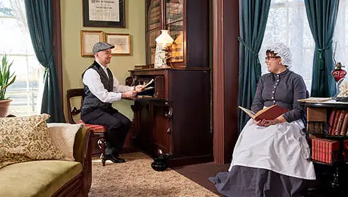IÀ l'intérieur du Bell Homestead, un homme et une femme vêtus de vêtements de 1874 se sourient en lisant, assis sur des chaises.