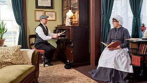 Inside the Bell Homestead, a man and woman dressed in clothes from 1874 smile at each other while sitting in chairs and reading