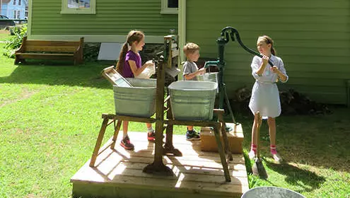 Drei Kinder füllen Metalleimer mit einer Wasserpumpe im alten Stil auf einer Holzplattform.