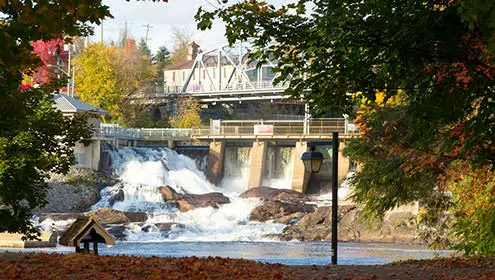 Bracebridge Falls desde el otro lado del río en la parte inferior de las cataratas. Hay hojas caídas de colores brillantes en el suelo y sobre las cataratas está el Puente de Plata.