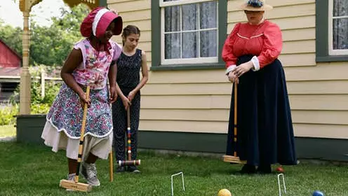 One woman and two young girls dressed in pioneer clothing play croquet next to historic house.