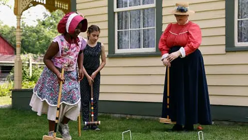 Une femme et deux jeunes filles en habits de pionnier jouent au croquet à côté d'une maison historique.