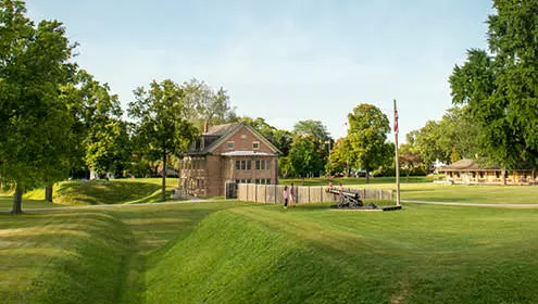 A view of Fort Malden National Historic site from across the grass. It is a a sunny day, people are walking in the distance. 