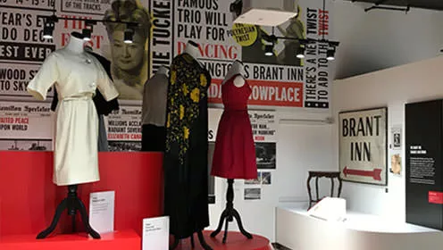 An indoor clothing exhibit at the Joseph Brant Museum. The room is filled with  mannequins styled with various  dresses. Pictures of women line the back wall along with quotes and titles from the exhibit.