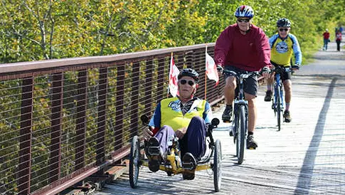 Cyclists crossing over a bridge on the Trans Canada Trail on a sunny day. There are pedestrians walking on the gravel trail in the background surrounded by lush greenery.