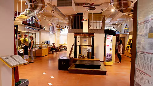 An interior view of the North Bay Museum. There information boards along the walls, a dislpay case to the left and stairs in the middle of the room. There are model planes hanging from the ceiling.