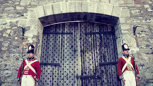 Deux soldats vêtus d'uniformes de la guerre de 1812 se tiennent de chaque côté d'une grande porte en bois menant à un grand fort en pierres.