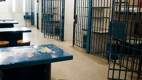 A hallway lined with open jail cells inside the Historic SDG Jail. On the left hand side are blue metal tables with stools.