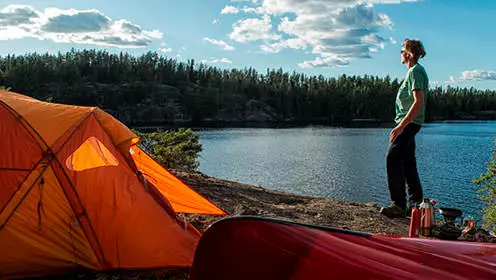 A man standing beside his tent and canoe on the shores of the lake on a sunny day. The distant shoreline is made up of rock cliffs.