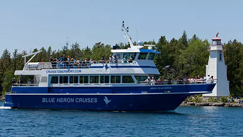 A large boat with people on the front and top deck sailing by a lighthouse. The boat has &quot;Blue Heron Cruises&quot; written on the side.