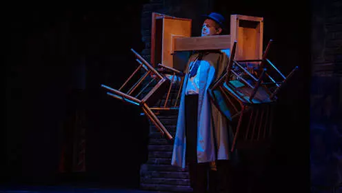 Ein Mann mit Trenchcoat und Hut steht mit blauem Scheinwerferlicht auf ihm. Er hält zwei Stühle in jeder Hand und balanciert ein Podest mit den Stühlen