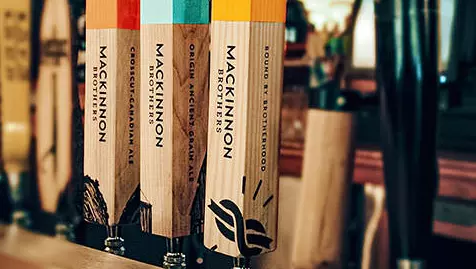 Tres grifos de cerveza de madera etiquetados con diferentes variedades de cerveza Mackinnon Brothers. Crosscut Canadian Ale, Origin Anceint Grain Ale y Bound By Brotherhood con etiqueta roja, azul o amarilla