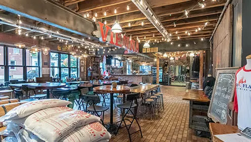 개방형 천장과 스트링 조명이 있는 넓은 방입니다. 다양한 크기의 테이블과 티셔츠를 입은 마네킹과 보리가 든 가방이 방을 가득 채웁니다. VIENNA라는 단어는 천장에 가까운 나무 기둥을 따라 수평으로 표시됩니다.