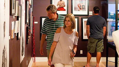 A man and a woman looking at the art displayed on the wall of an art gallery. Another man is shown in the background looking at art.