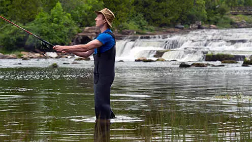 A man wearing waders on the shores of a lake is casting a fishing rod. There is a waterfall in the distance.