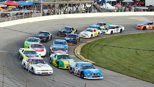 Fifteen race cars speeding around the bend of an oval racetrack. There are tents in the background as well as a spectators standing on a wooden observation deck.