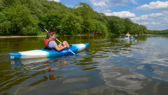 Femme pagayant en kayak sur une rivière bordée d'arbres et qui suit un second kayak.