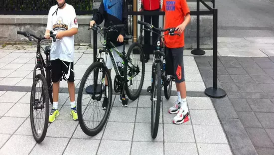 Trois jeunes enfants qui sourient et qui marchent à côté de leur vélo à côté d'une auto-patrouille de la GRC stationnée entre deux colonnes de pierre.